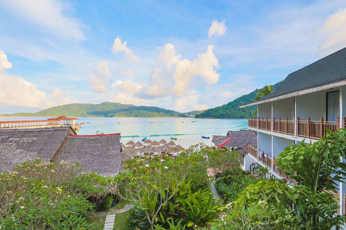 A beachfront hotel with a view of the sea, a thatched-roof area, and lush greenery.