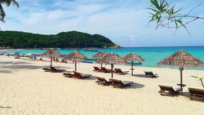 Ein Strand mit Liegestühlen und Reetdachschirmen, ein klares blaues Meer und ein bewaldeter Hügel im Hintergrund.