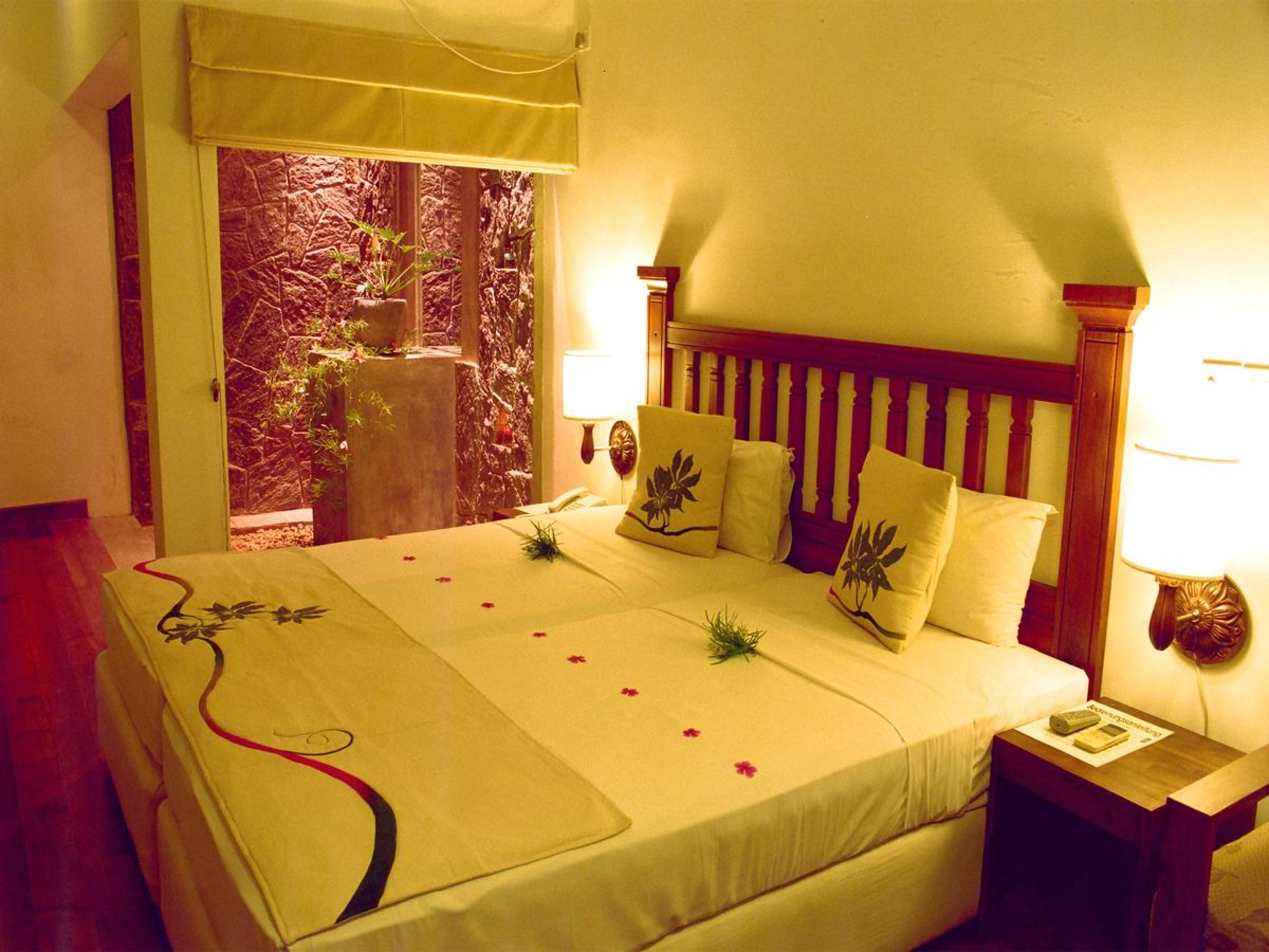 A hotel room with a bed, a nightstand, a lamp, and a view of a garden through a window.