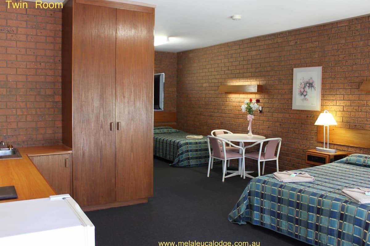 A twin room with a brick wall, wooden wardrobe, two beds with blue checkered bedding, a small table with chairs, a lamp, and a framed picture on the wall.