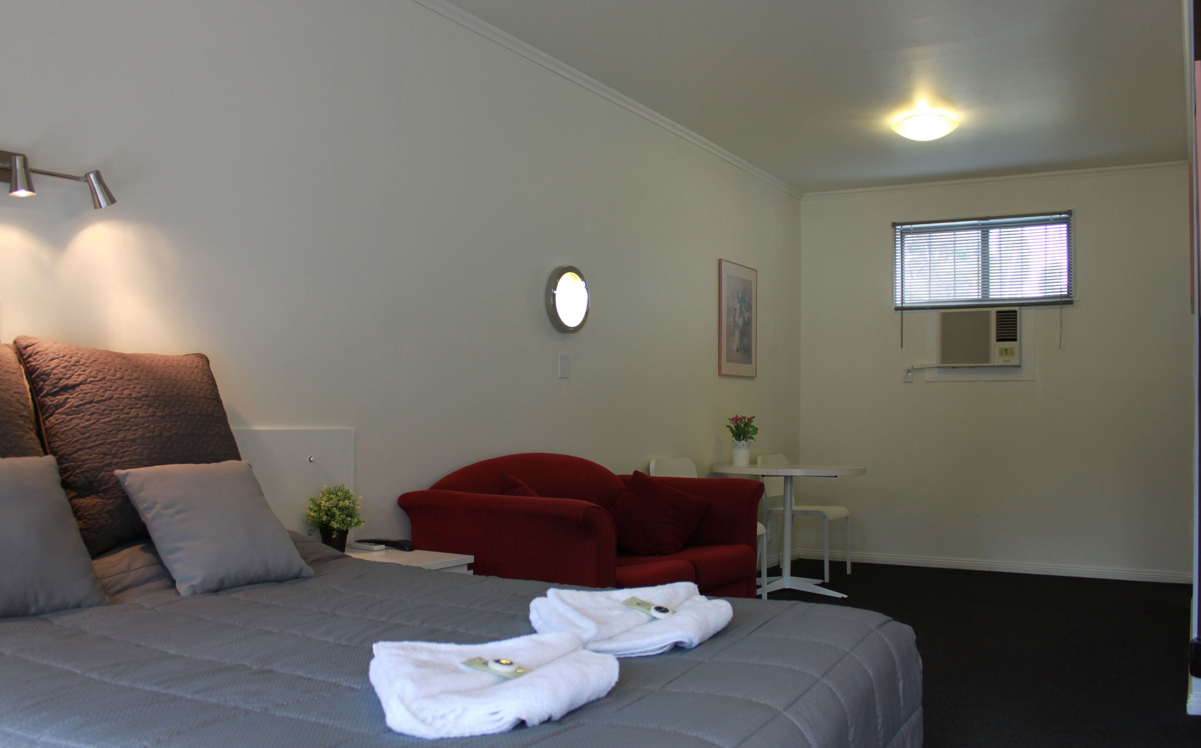 A hotel room with a bed, a red armchair, a small table with a plant, and a window with blinds.
