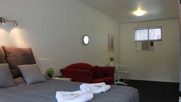 A hotel room with a bed, a red armchair, a small table with a plant, and a window with blinds.