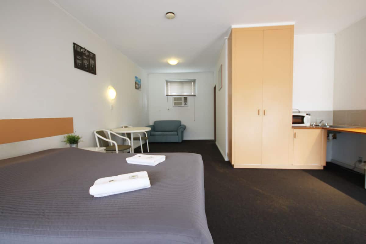 A hotel room with a bed, a desk, a chair, a kitchenette, and a small window.