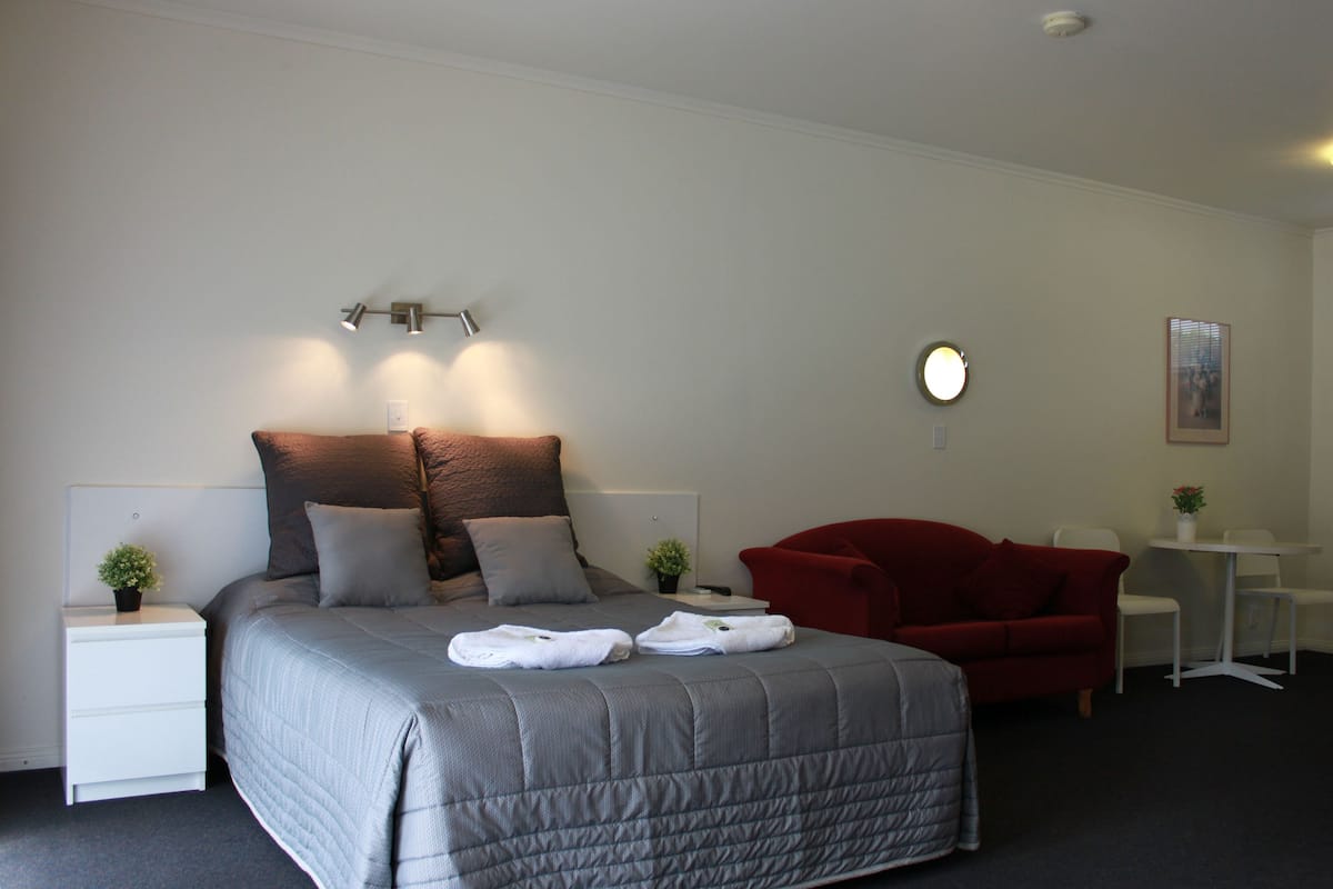 A neatly arranged bedroom with a bed, a red armchair, a small table, and a wall-mounted light fixture.