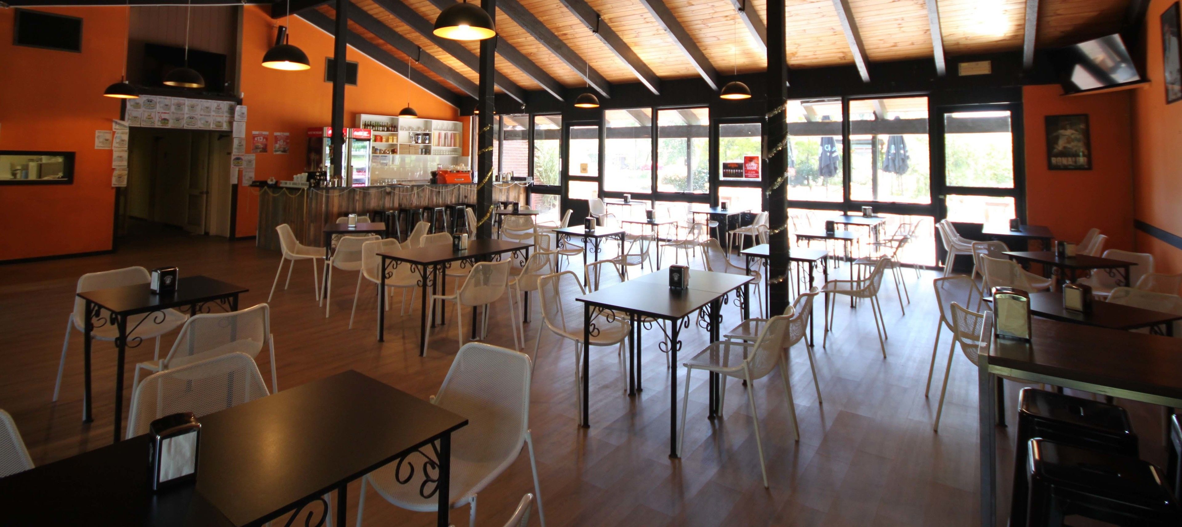 A spacious indoor dining area with multiple tables and chairs, a bar area with shelves of bottles, and large windows allowing natural light.