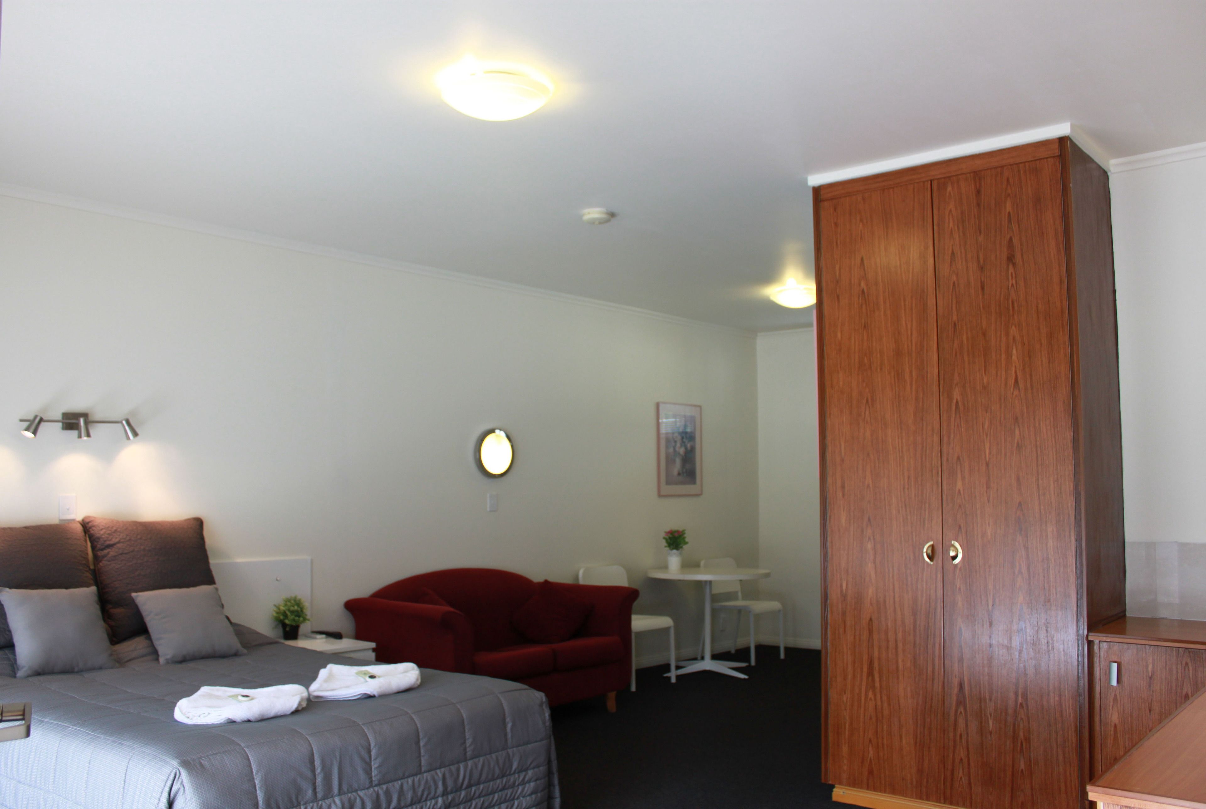 A hotel room with a bed, a red sofa, a wooden wardrobe, a small table with chairs, and a wall-mounted light fixture.