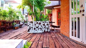 An outdoor wooden deck with a white decorative partition, surrounded by potted plants and a brick building with a glass door. - Hy Resort (Chonburi)