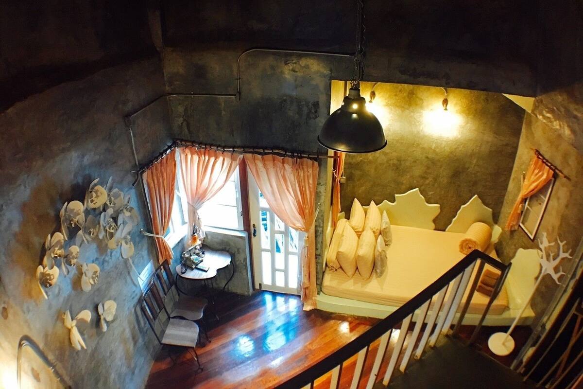 Una habitación con escalera de madera, cama con cabecera original, mesa con sillas y ventana con cortinas.