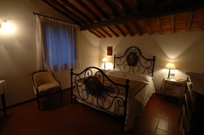 A bedroom with a four-poster bed, a chair, a bedside table with a lamp, and a window with curtains. - Albergo Il Colombaio (Castellina in Chianti)