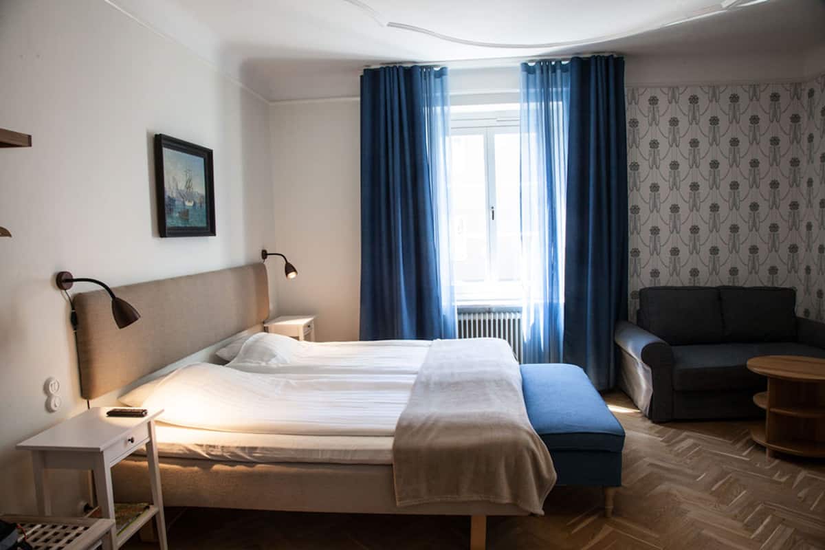 A hotel room with a bed, bedside tables, a lamp, a sofa, and a window with curtains.