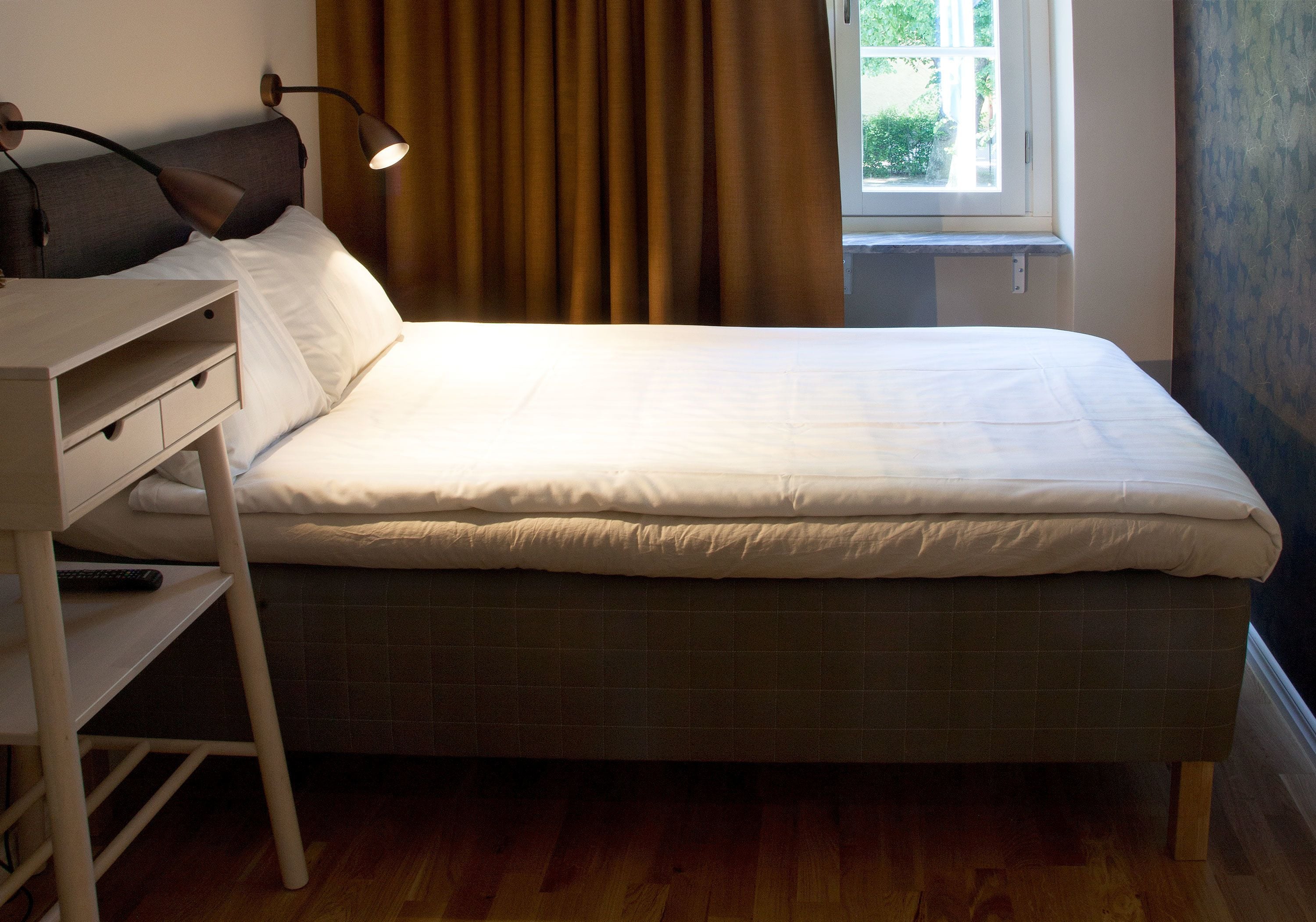 A neatly made bed with a headboard, a bedside table with a lamp, and a window with curtains.