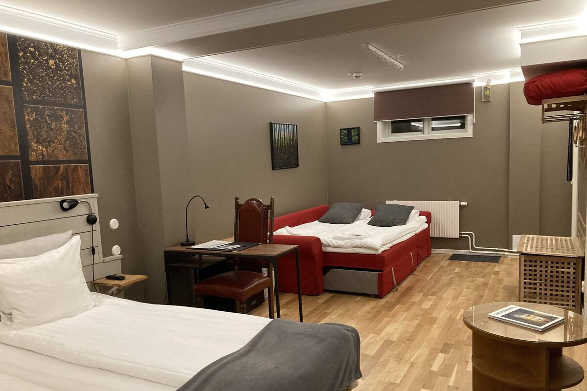 A hotel room with a bed, a desk, a chair, a red sofa, and a small table with a magazine.