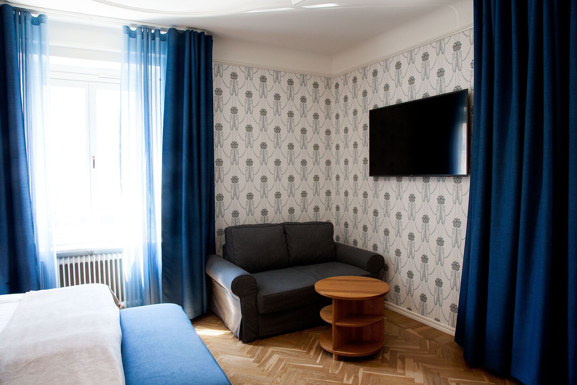 A hotel room with a sofa, a small wooden side table, a TV mounted on the wall, and patterned wallpaper.