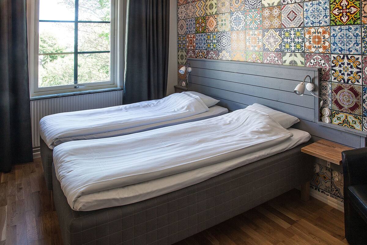 A room with two beds, a window with curtains, a tiled wall, and a wooden floor.