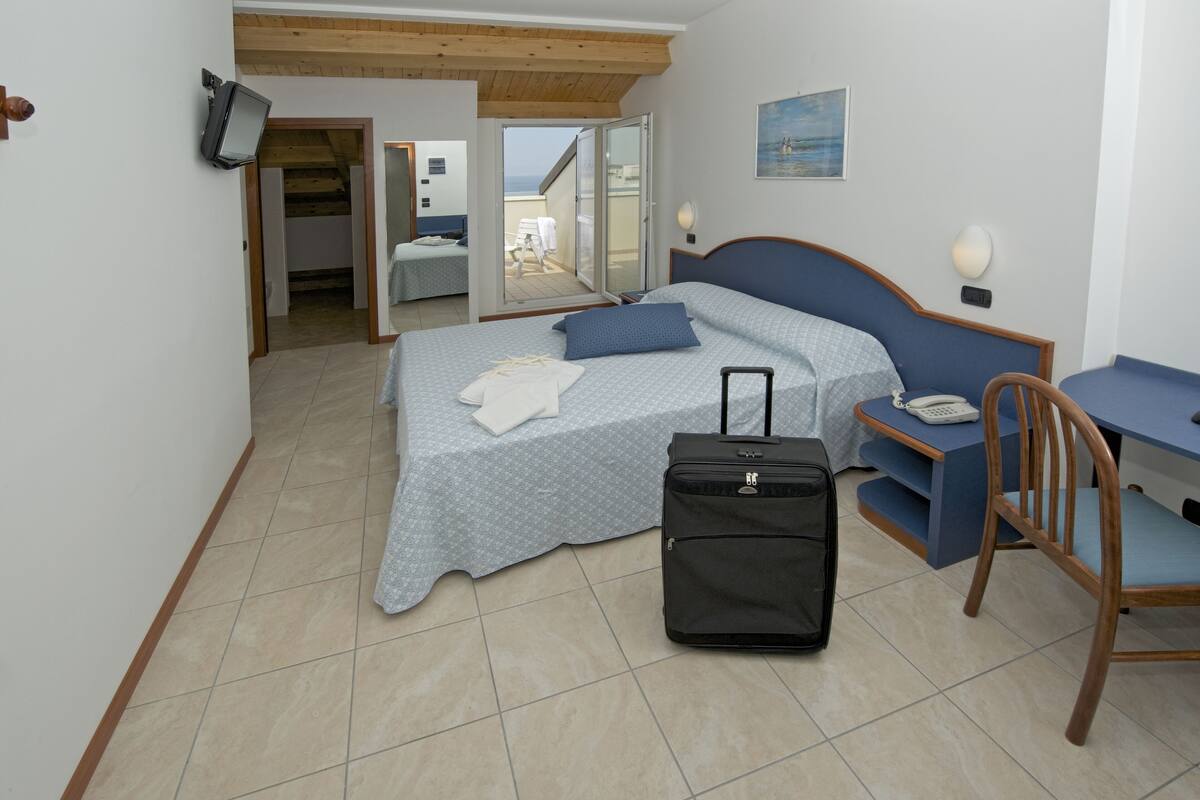 A bedroom with a bed, a desk, a chair, a suitcase, and a balcony with a view.