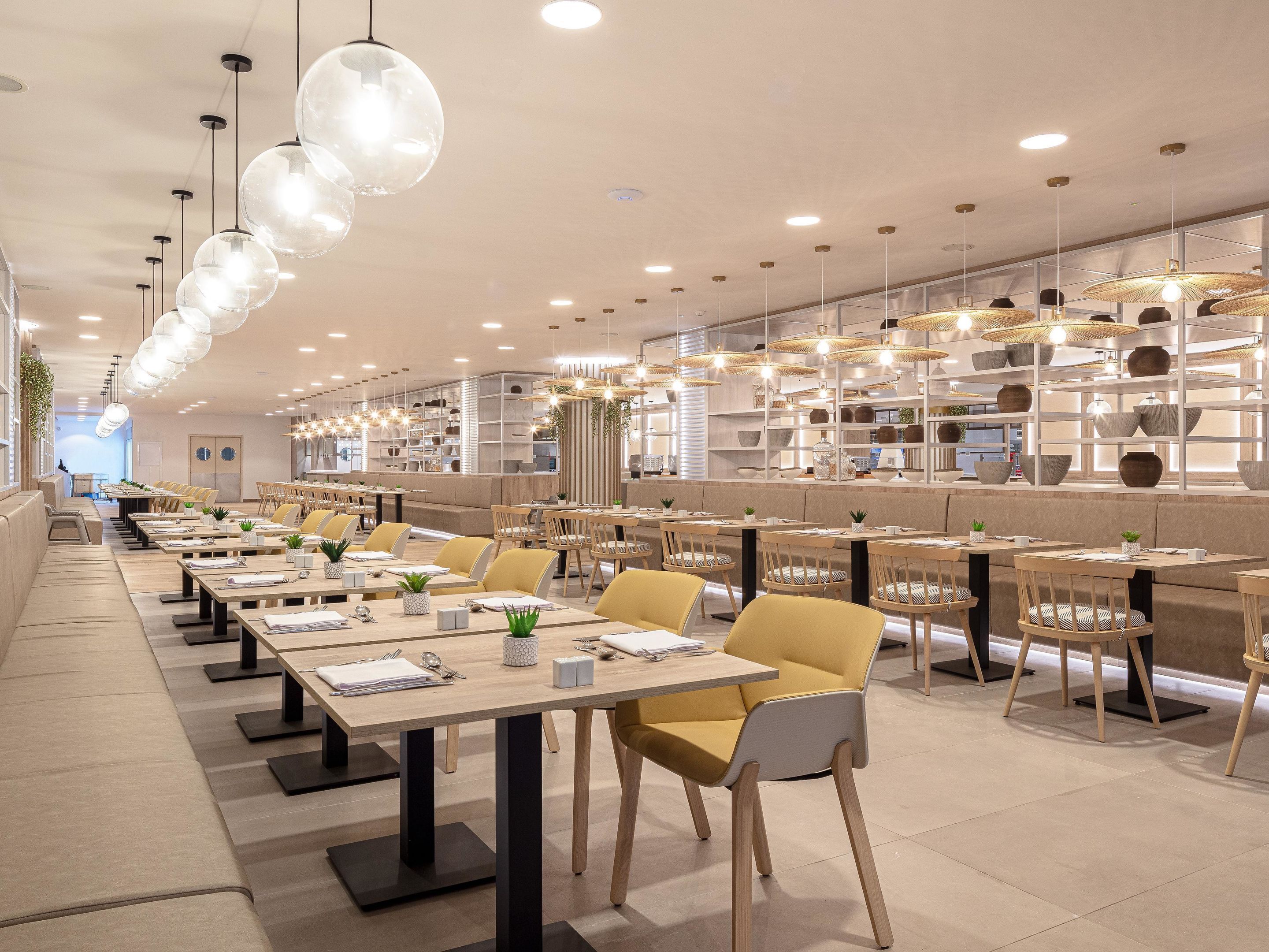Un restaurante moderno con mesas largas compartidas, sillas amarillas e iluminación colgante.