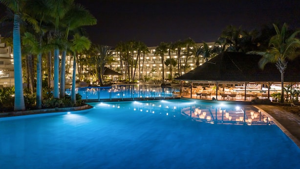 A nighttime swimming pool with palm trees and a building in the background. - Hotel Maspalomas Princess-All Inclusive (San Bartolomé de Tirajana)