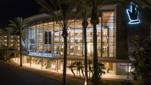 A modern hotel with a glass facade, palm trees, and a neon sign. - Hotel Maspalomas Princess-All Inclusive (San Bartolomé de Tirajana)