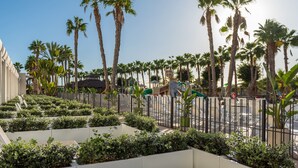 A landscaped area with palm trees, a fenced section, and a building in the background. - Hotel Maspalomas Princess-All Inclusive (San Bartolomé de Tirajana)