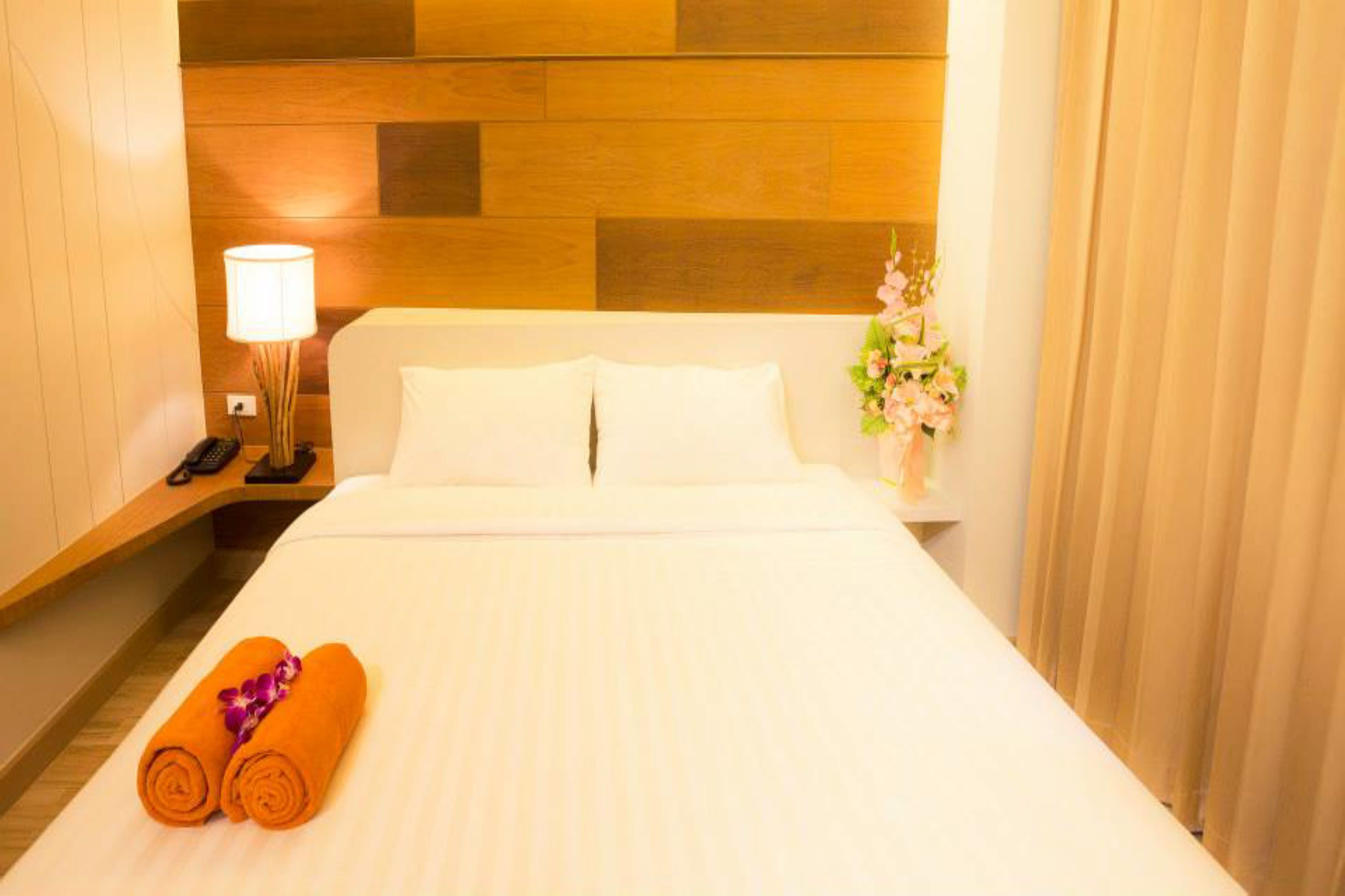 A hotel room with a wooden headboard, a bedside lamp, a vase with flowers, and rolled towels on the bed.