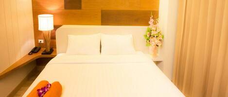 A hotel room with a wooden headboard, a bedside lamp, a vase with flowers, and rolled towels on the bed.