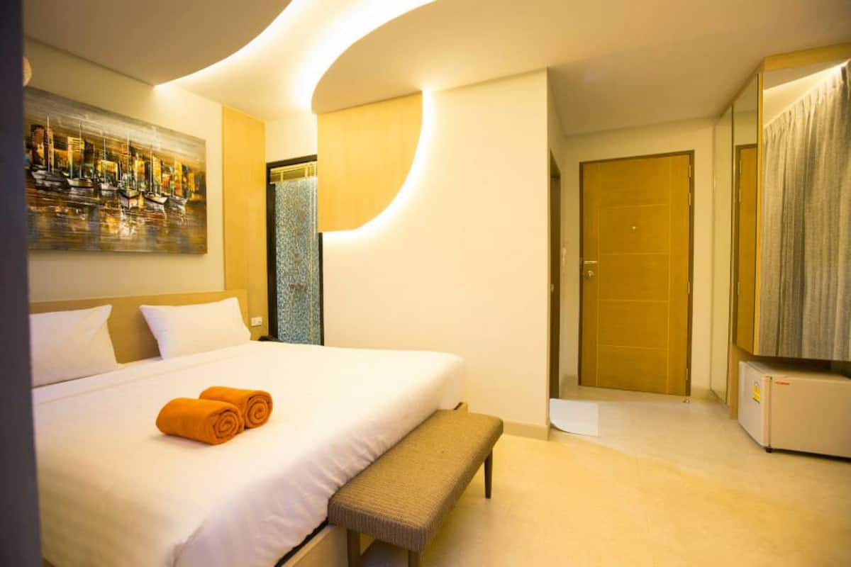 A hotel room with a large bed, a bench, a painting, a mirror, and a door.