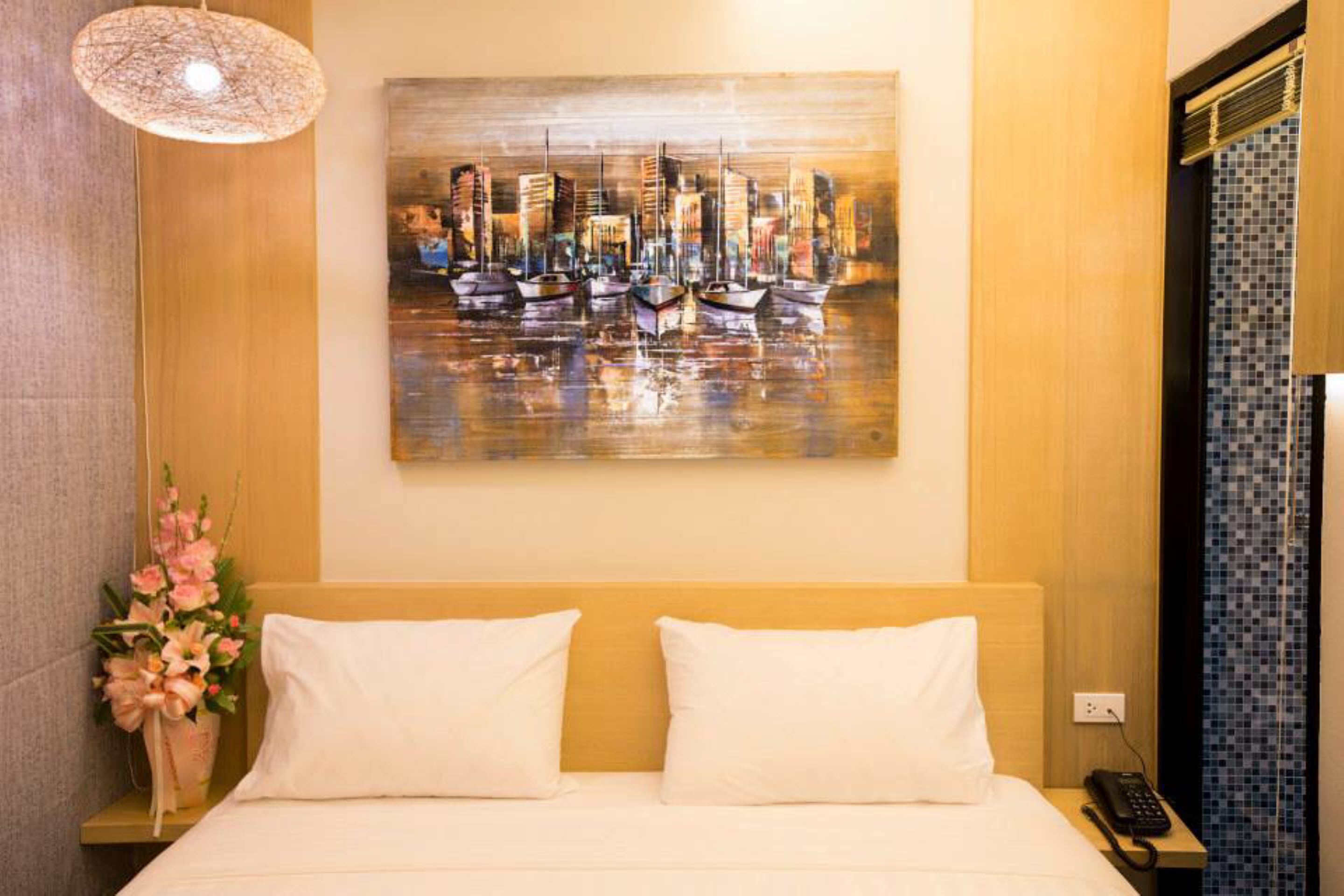 A hotel room with a bed, a painting of boats on water, a vase with flowers, and a telephone on the nightstand.