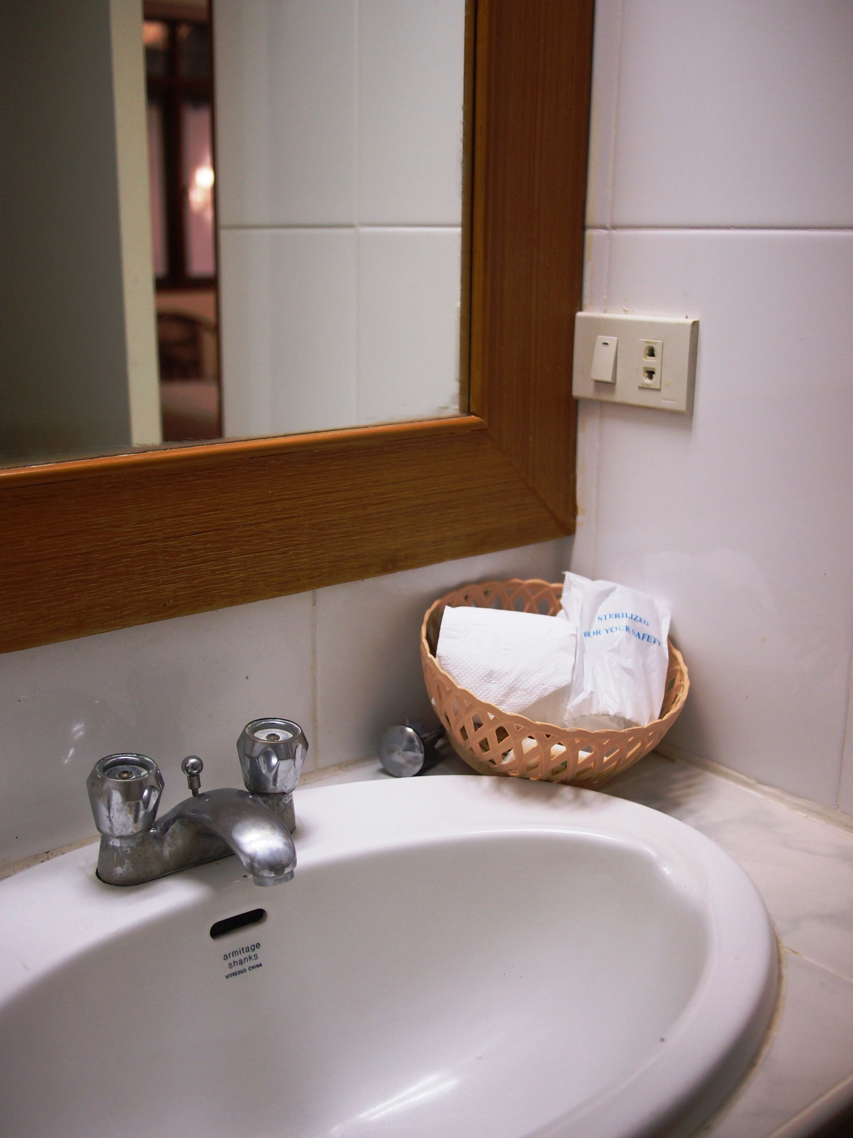 A bathroom sink with a mirror, a wooden-framed mirror, a basket with a tissue, and a faucet.