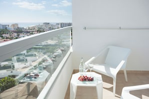 A balcony with a glass railing, a white chair, a small table with a bottle and glasses, and a view of a cityscape.