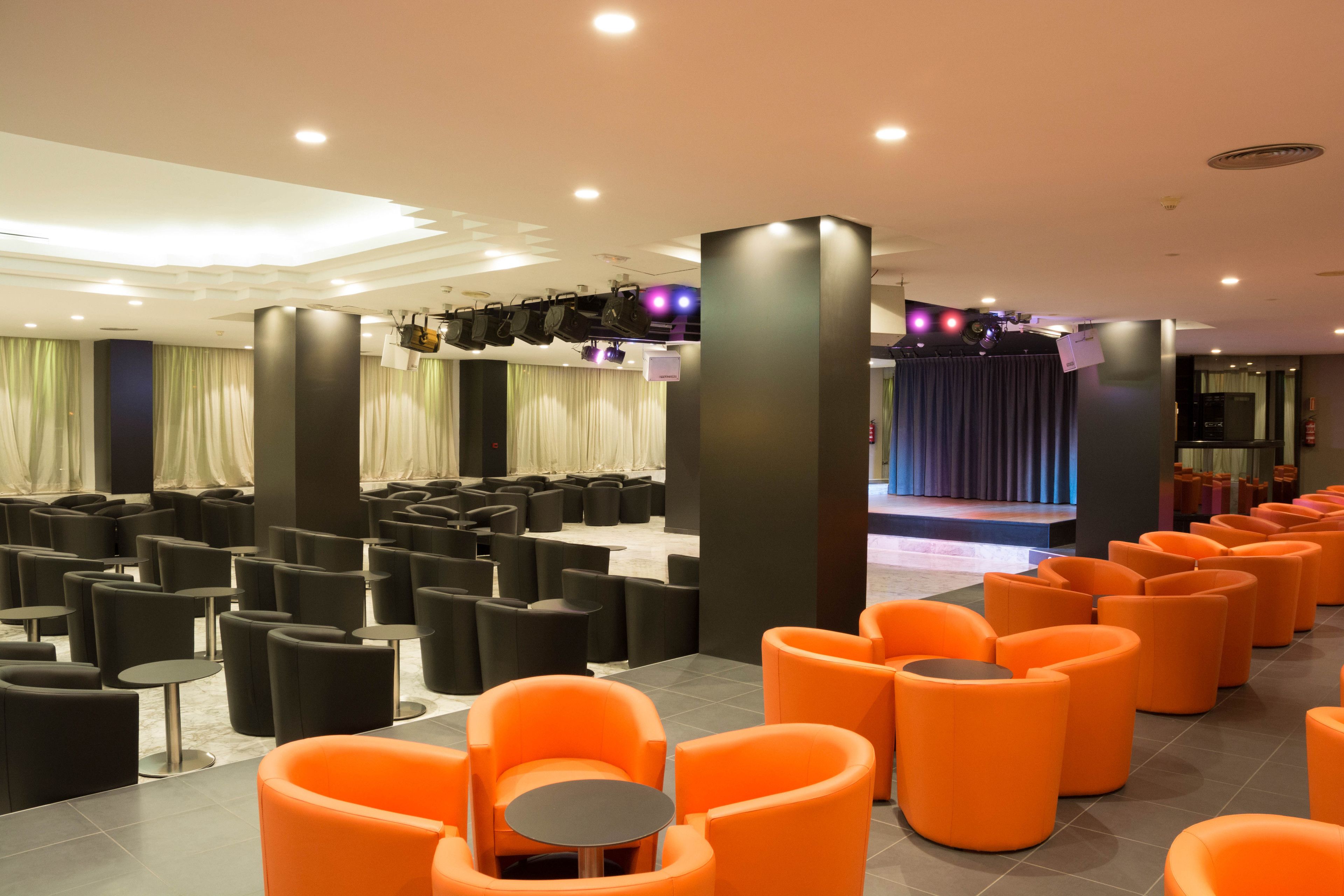 Un moderno vestíbulo de hotel con filas de asientos negros y naranja, un escenario e iluminación empotrada en el techo.