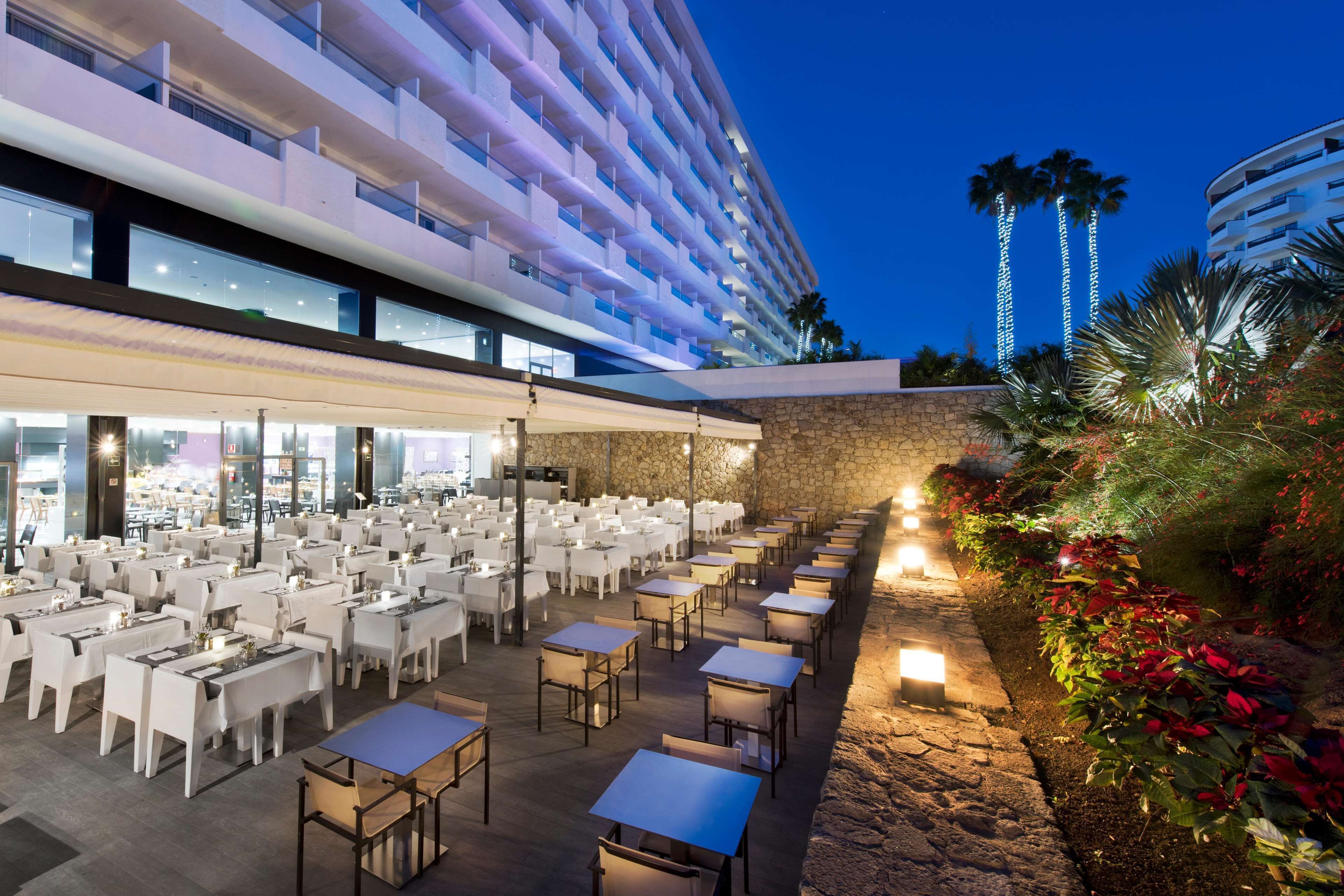 An outdoor dining area with tables and chairs, a stone wall, and a building with balconies in the background.