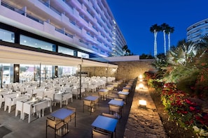An outdoor dining area with tables and chairs, a stone wall, and a building with balconies in the background. - Hotel Gran Canaria Princess - Adults Only (+16) (San Bartolomé de Tirajana)