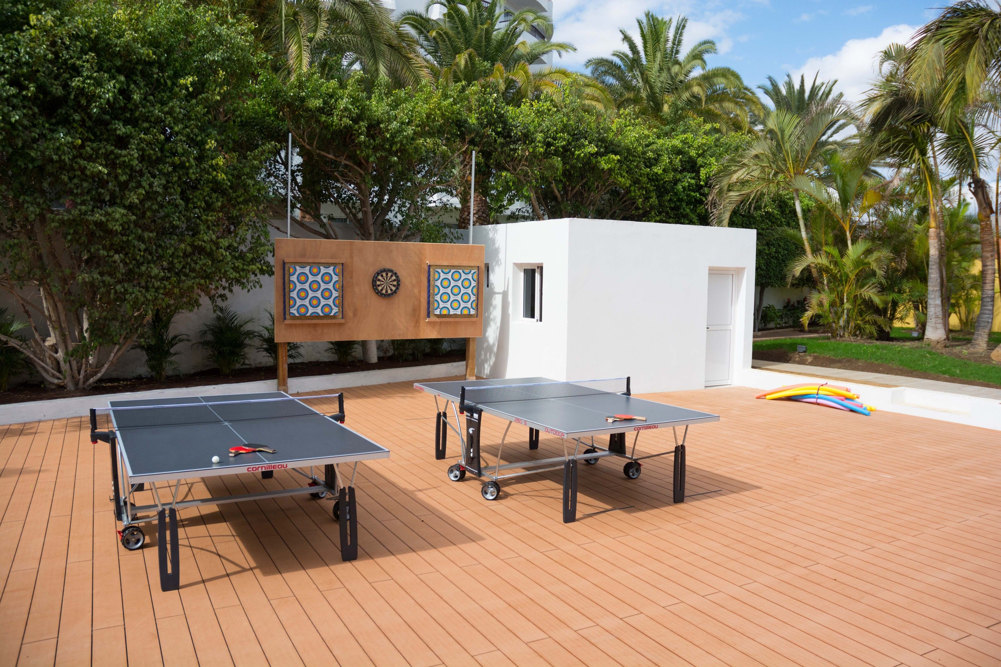 Two ping pong tables on a wooden deck with palm trees and a small white building in the background.