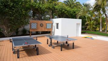 Due tavoli da ping pong su un pontile in legno, con palme e un piccolo edificio bianco sullo sfondo.