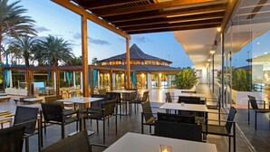 An outdoor dining area with tables and chairs, a covered seating area, and a building with a thatched roof in the background. - Hotel Gran Canaria Princess - Adults Only (+16) (San Bartolomé de Tirajana)