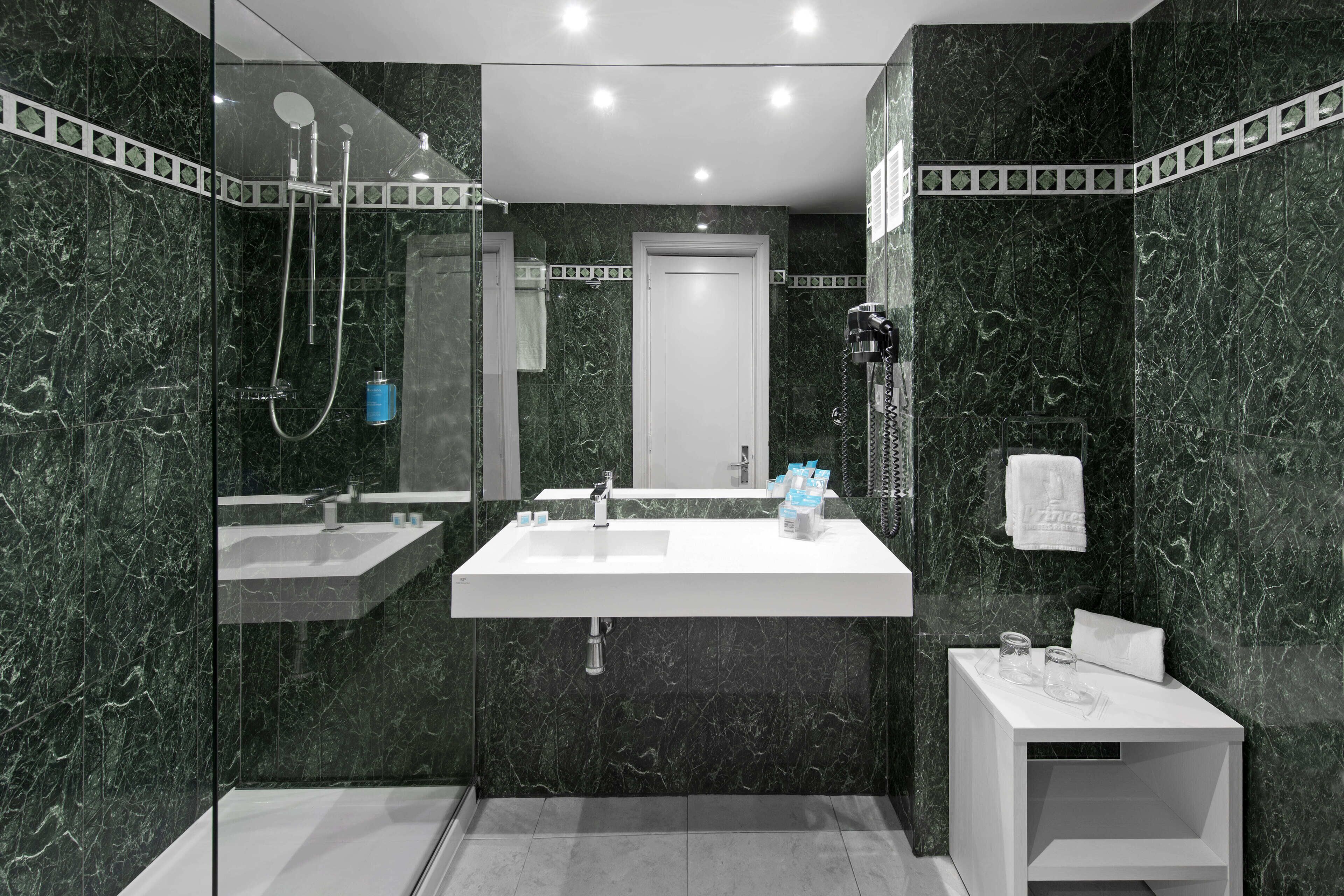 A modern bathroom with a large mirror, two sinks, a shower, and a small bedside table with a towel.