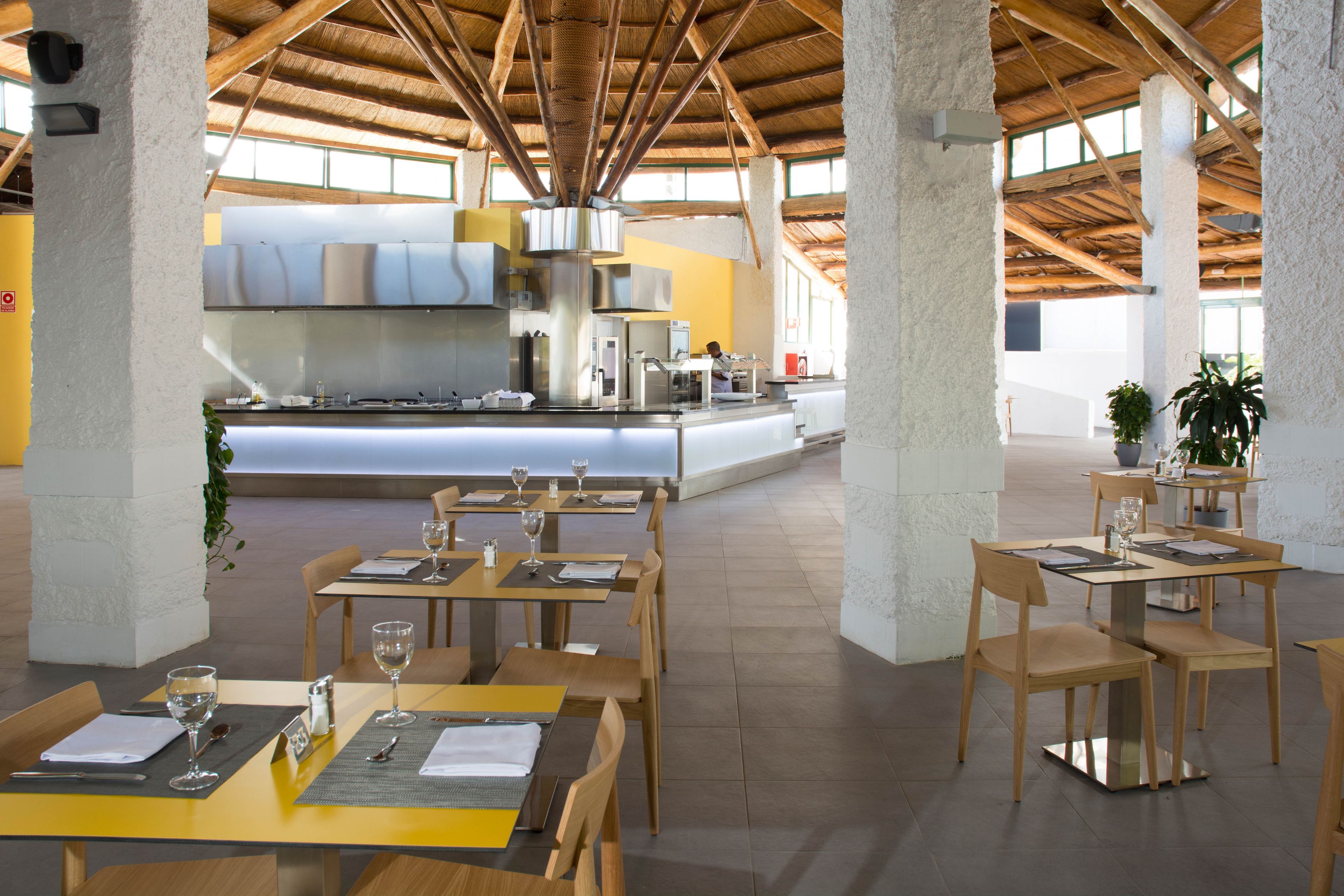 Un comedor amplio con mesas y sillas de madera, una zona de cocina central y grandes ventanales que dejan entrar luz natural.