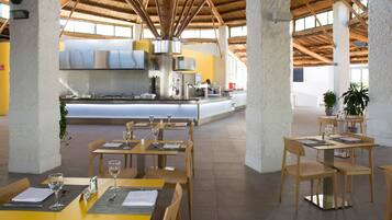 Un comedor amplio con mesas y sillas de madera, una zona de cocina central y grandes ventanales que dejan entrar luz natural.