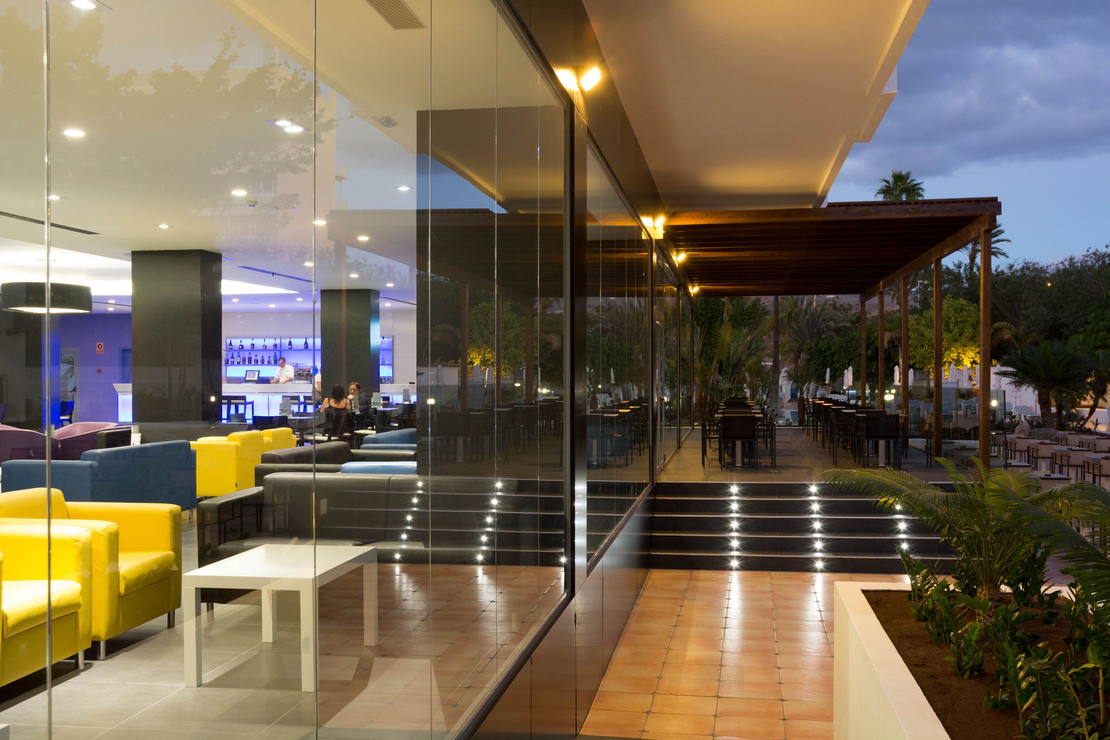 Un espace extérieur moderne, doté de murs en verre, d’un bar et d’un mobilier composé de sièges aux couleurs jaune et gris.