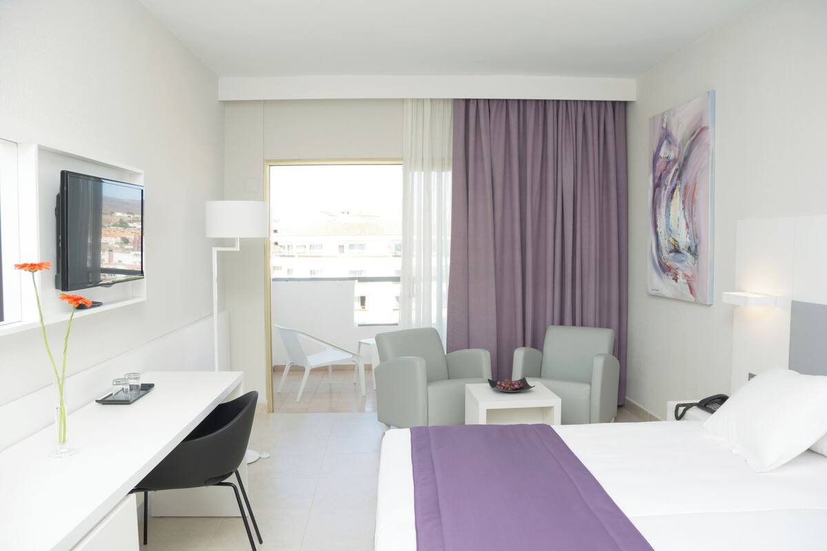 A hotel room with a bed, a desk, a chair, a small table, a TV, and a window with curtains.