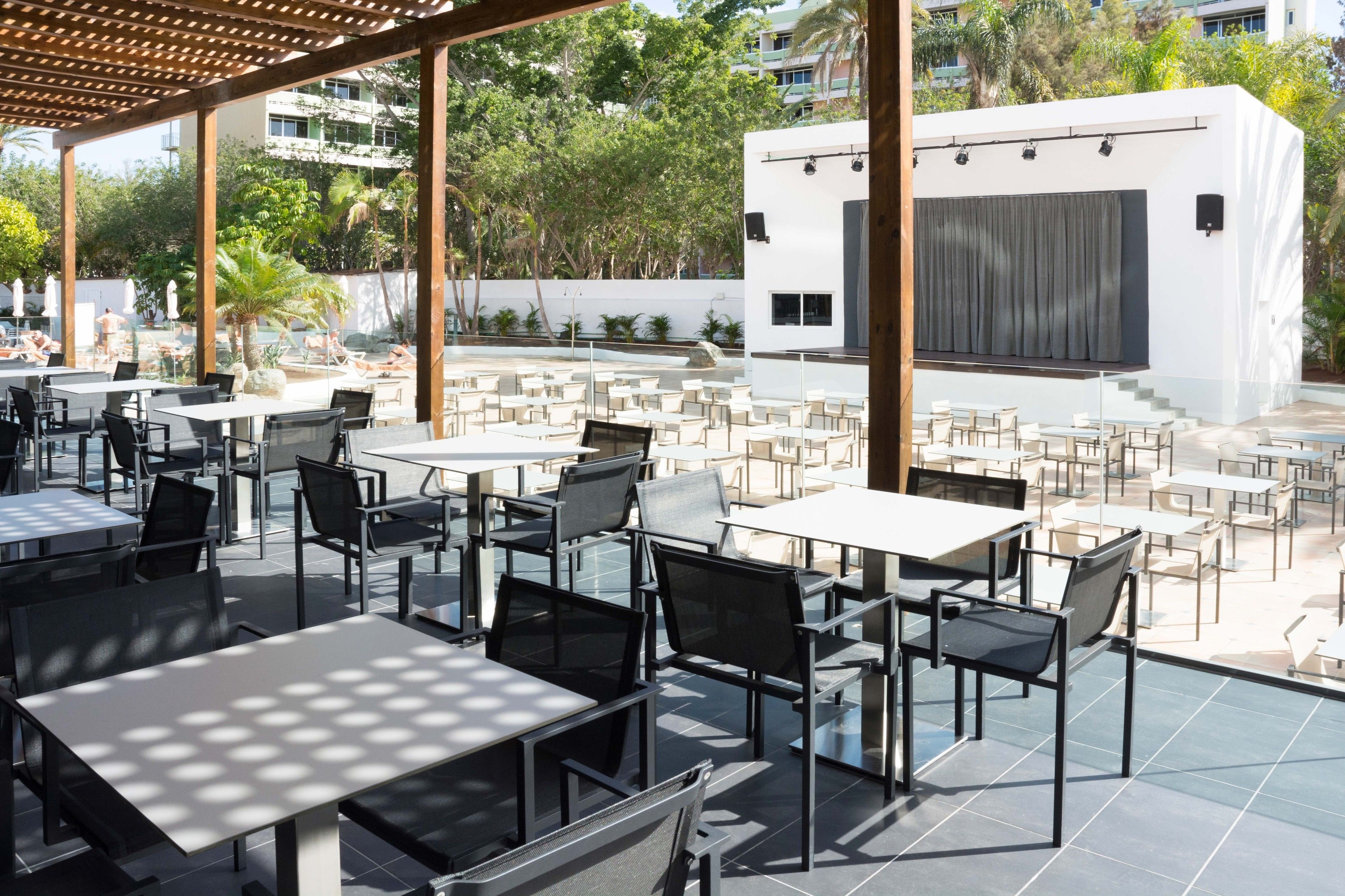 Un área de comedor al aire libre con varias mesas y sillas, un escenario y una piscina al fondo.
