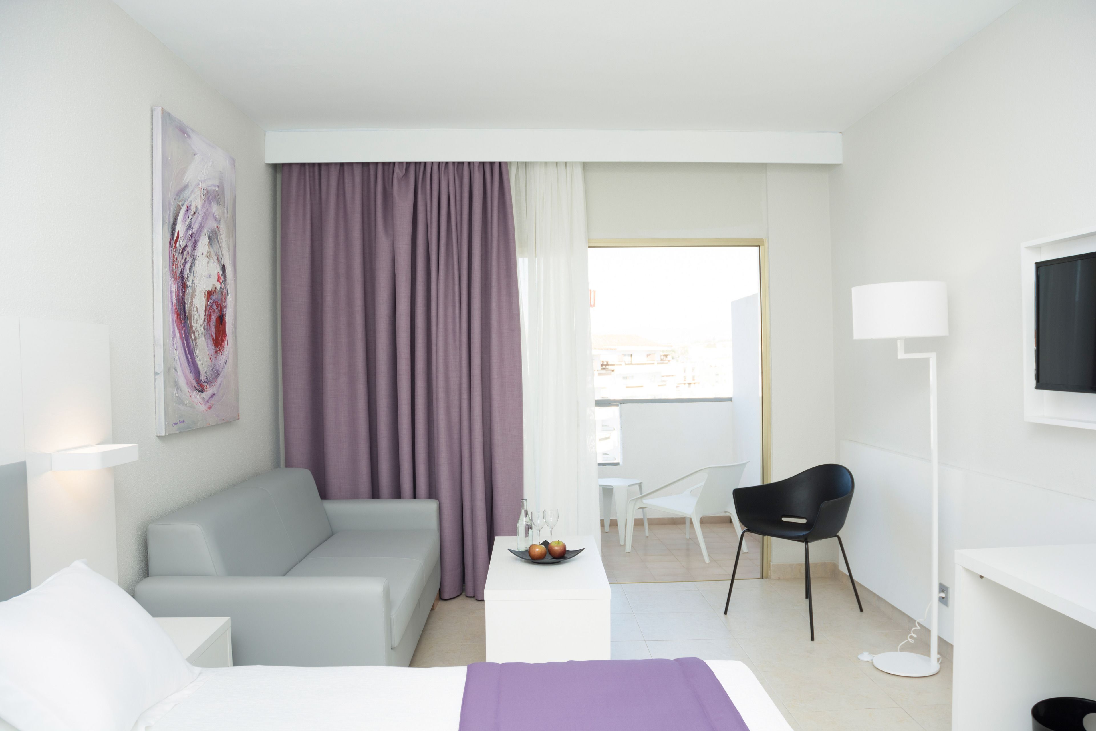 A modern hotel room with a bed, a grey sofa, a small dining table with fruit, a black chair, a white desk, and a large window with curtains.