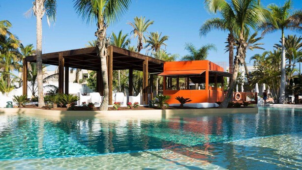 A clear pool with a wooden deck and lounge chairs, surrounded by palm trees and an orange building. - Hotel Gran Canaria Princess - Adults Only (+16) (San Bartolomé de Tirajana)