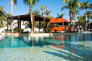 Ein klares Schwimmbecken mit Holzterrasse und Liegestühlen, umgeben von Palmen und einem orangen Gebäude.