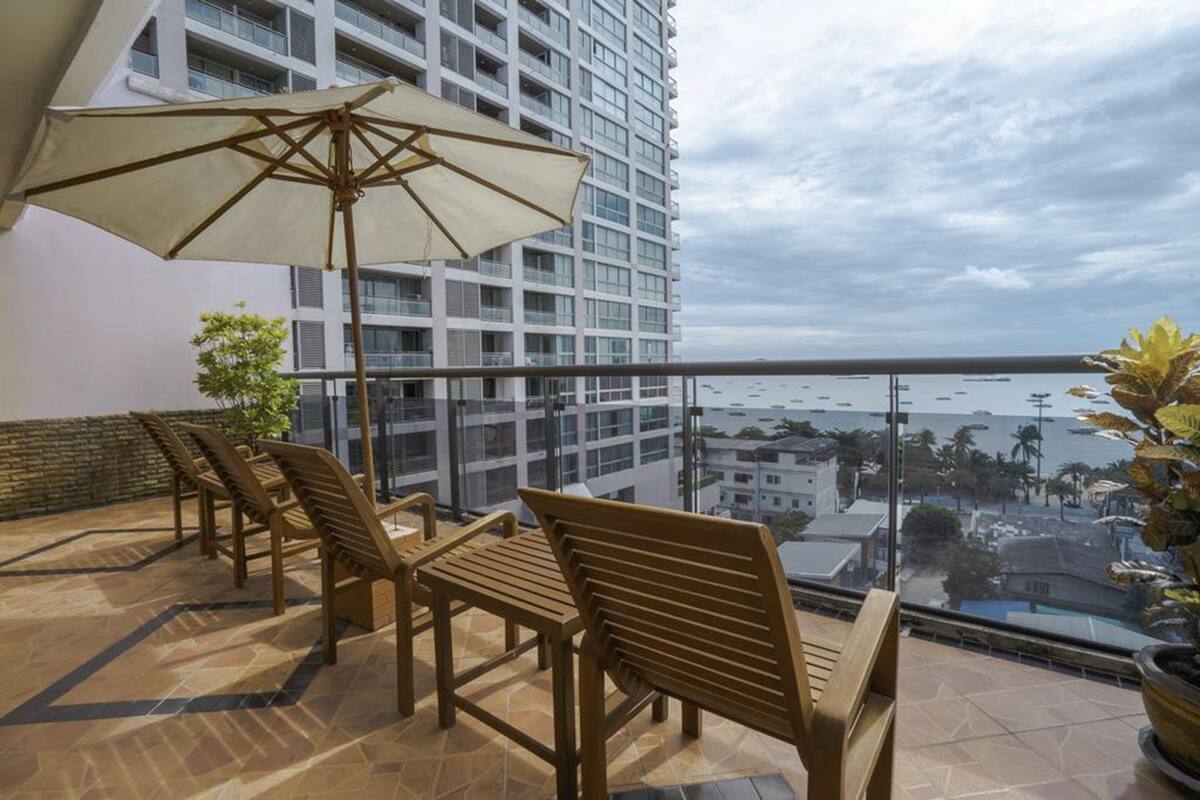 A balcony with wooden chairs and a large umbrella, overlooking a cityscape and the sea.