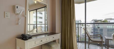 A hotel room with a vanity, a telephone, a mirror, a window with a view of buildings, and a balcony with a chair.