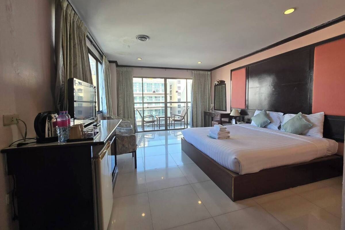 A hotel room with a bed, a desk with a chair, a TV, and a balcony with a view of the city.