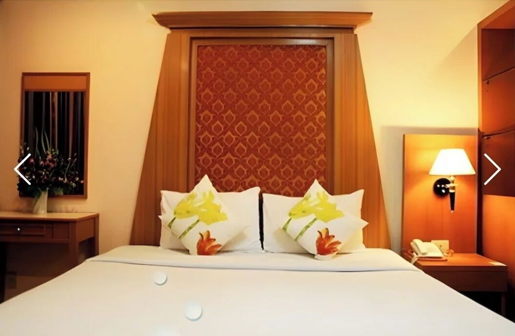 A hotel room with a large bed, two pillows with floral designs, a bedside table with a lamp, and a mirror on the wall.