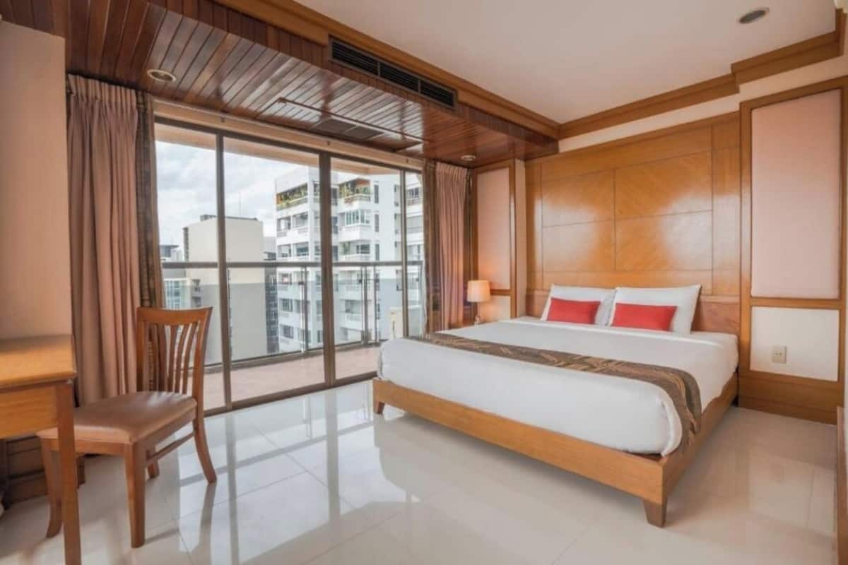 A hotel room with a large bed, a desk, and a chair. There is a large window with curtains, and the room has a wooden ceiling.