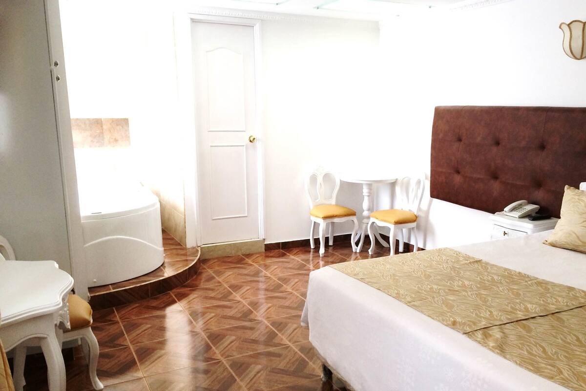A hotel room with a bed, a small round table with two chairs, a white door, and a window.